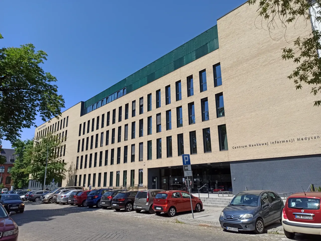 Pięciokondygnacyjny gmach z jasnej cegły z ciemnozielonym pasem najwyższej kondygnacji, z wypukłym napisem „Centrum Naukowej Informacji Medycznej” nad przeszkloną częścią parteru i samochodami zaparkowanymi wzdłuż brukowanej ulicy