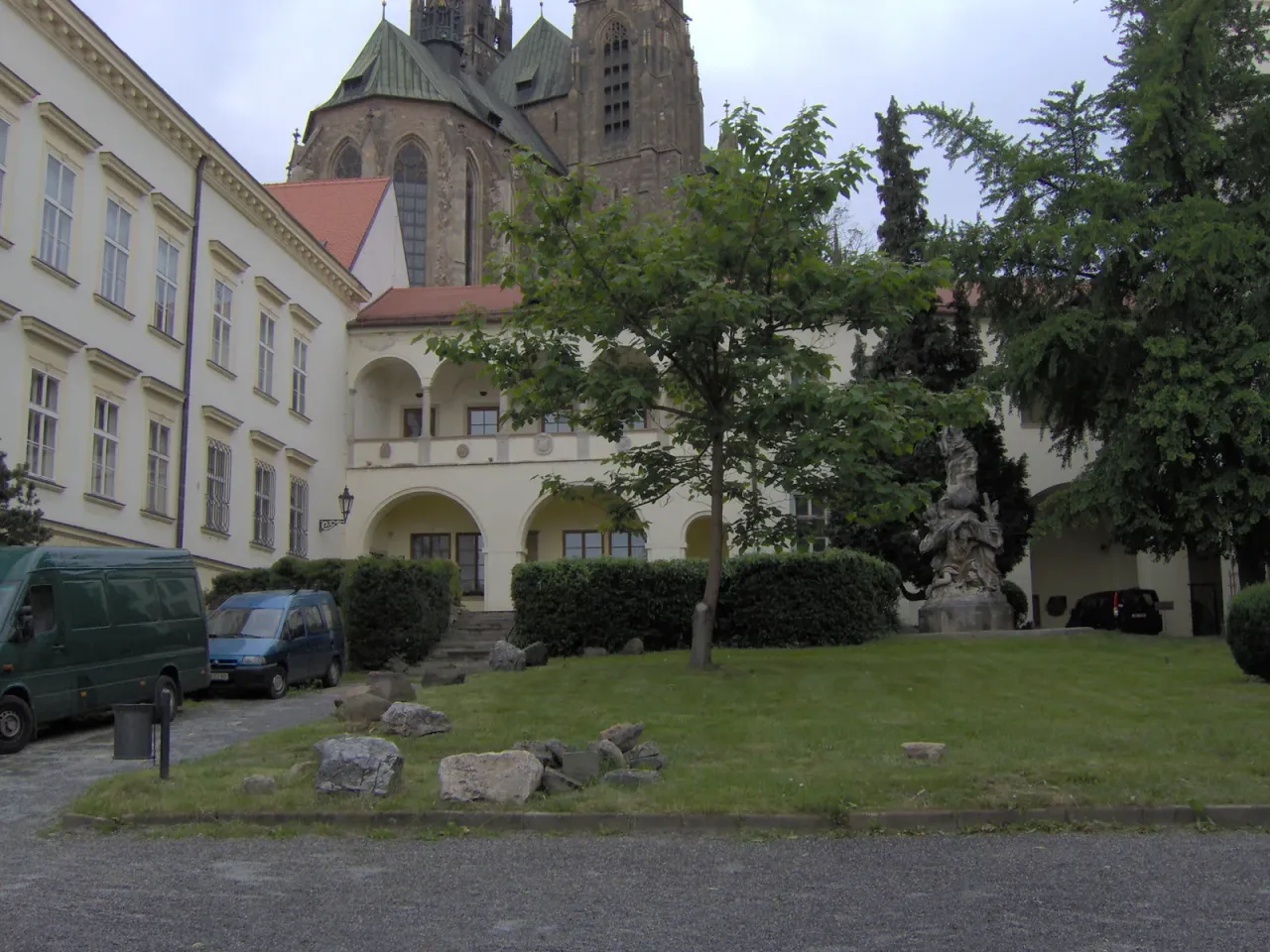 Trawiasty dziedziniec Biskupského dvora w Brnie: jasny budynek z renesansową loggią arkadową, kamienna grupa rzeźbiarska po prawej, młode drzewo pośrodku, w tle wieże katedry na Petrovie