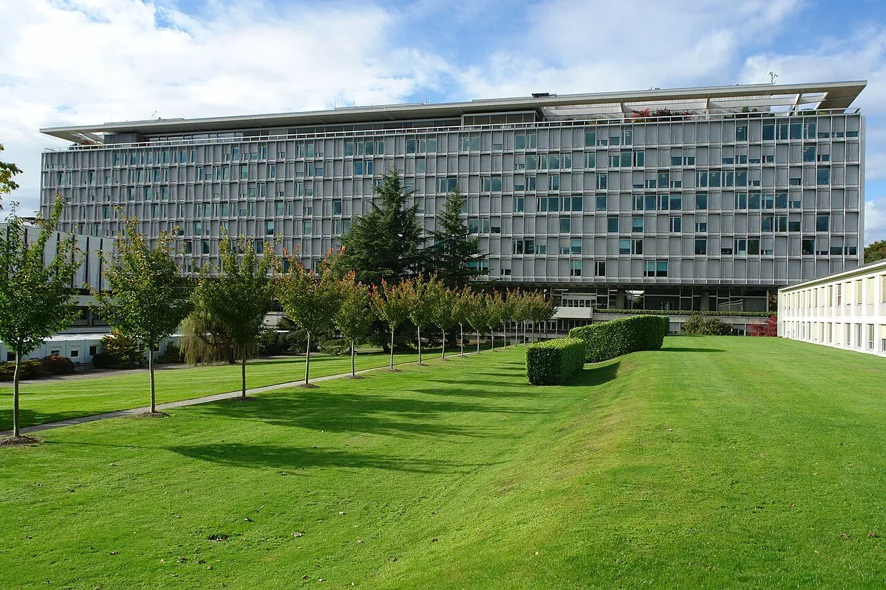 Długi modernistyczny gmach siedziby Światowej Organizacji Zdrowia w Genewie widziany od południa: szaro-srebrna elewacja z pionowymi żaluzjami i rzędami okien na kilku kondygnacjach, wysunięty płaski dach, a przed budynkiem rozległy trawnik, szpaler młodych drzew i przycięty żywopłot.